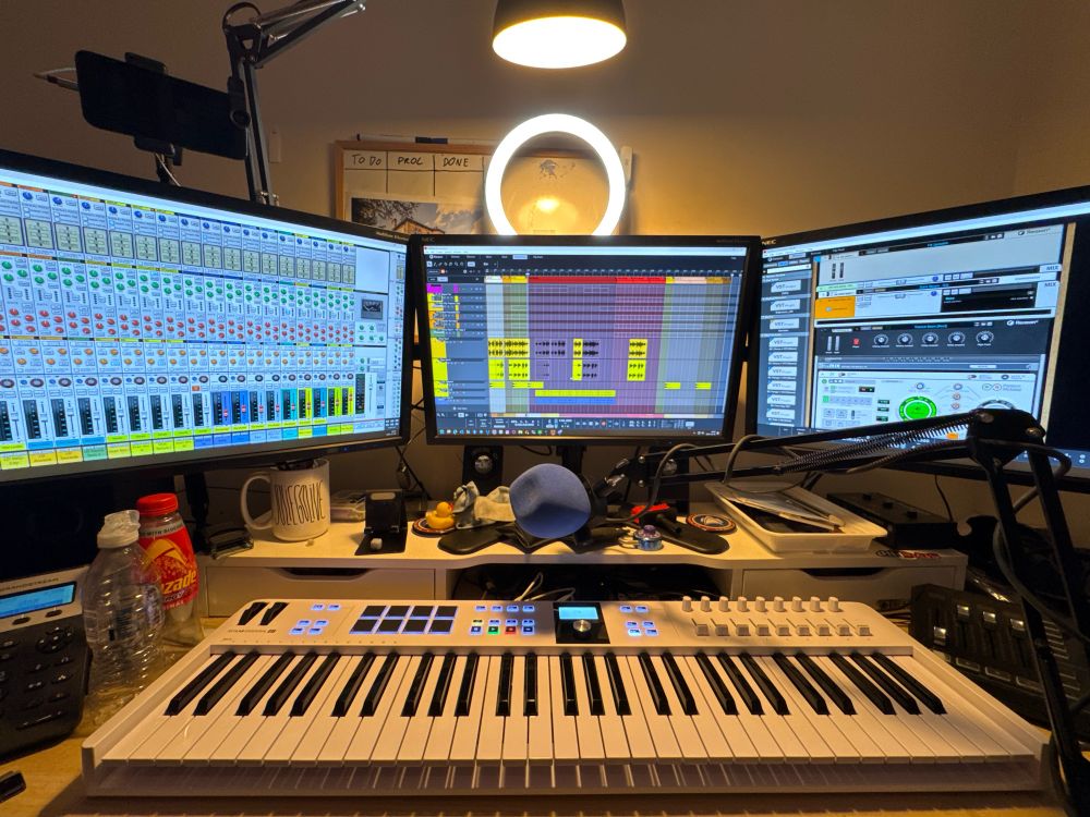 Wide shot of studio desk with 3 monitors showing sequencer/arranger, mixer, and device rack, mounted on top of slightly untidy desk riser with microphone and full length controller keyboard in foreground