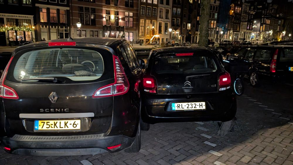 Two black cars with Dutch number plates. They're touching each other in a seemingly passive aggressive manner that encroaches on multiple parking spaces. 