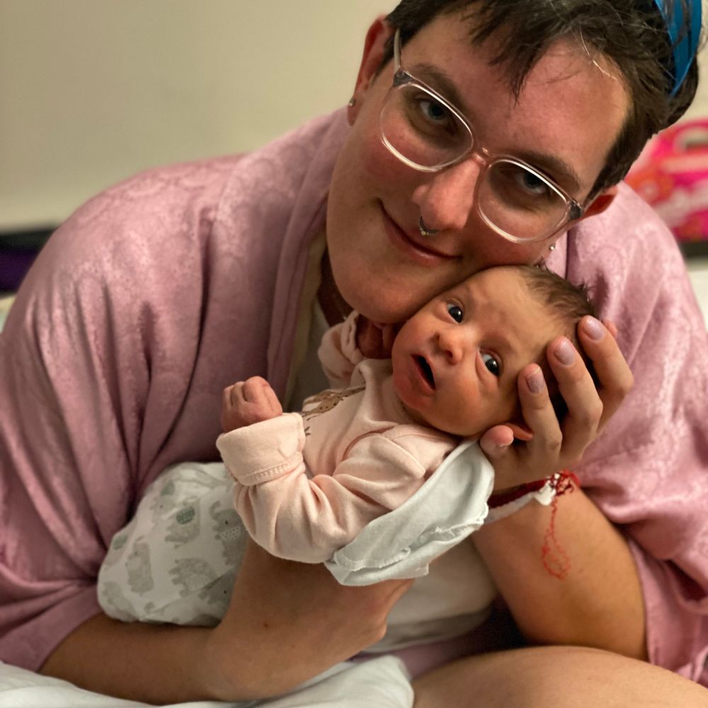 Tekla, a lovely white non-binary lesbian wearing a rose pink silk robe, cradles a newborn baby in their hands. The baby is alert and looking at the camera. She has a head of dark hair, wide open eyes, and is wearing a pink onesie 