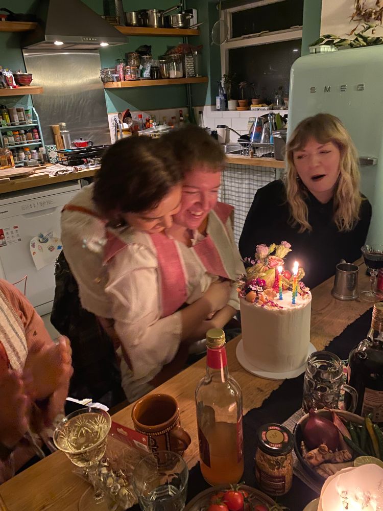 Hollie’s partner is hugging them as they smile at a giant white frosted cake. It has three rainbow coloured candles, roses and lots of fruit on top 