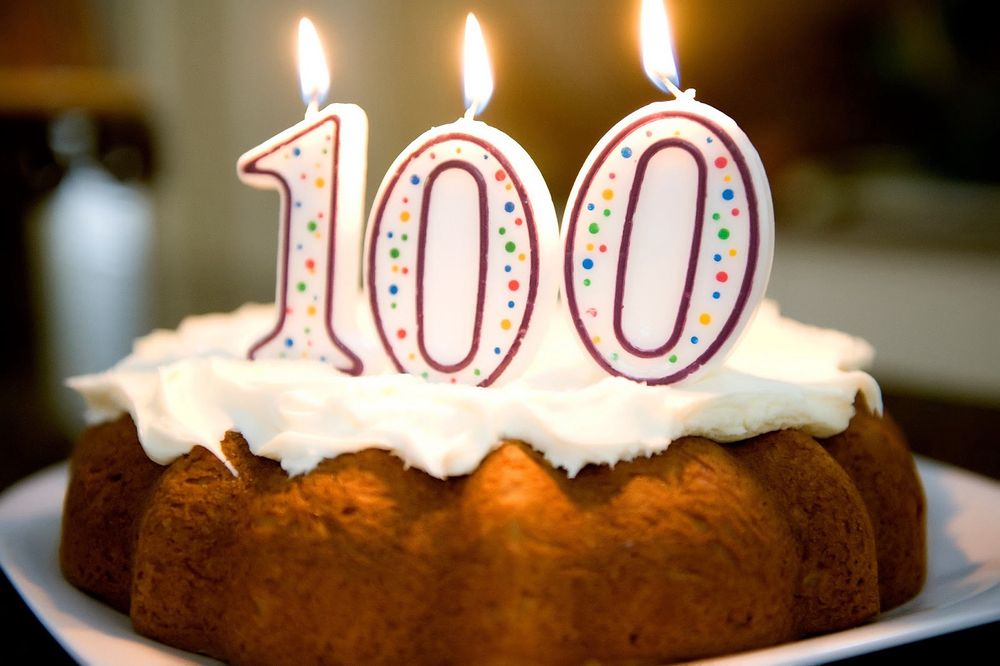 Bunt cake with three lit candles spelling out 100. 