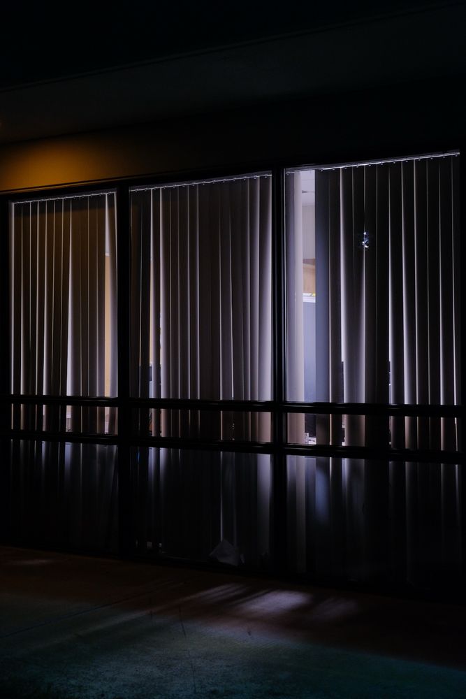 A photo of a dimly lit building exterior at night, featuring a row of large windows with vertical blinds. The blinds are mostly closed, but some are slightly parted, revealing glimpses of the interior. The scene is illuminated by a mix of warm and cool lighting, casting soft shadows on the ground