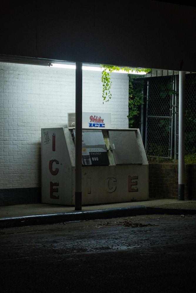 A photo of an old ice vending machine at night, illuminated by a harsh overhead fluorescent light. The machine is labeled "Holiday Ice" with large faded red letters spelling "ICE" on its side, with some small Sharpie graffiti in the top corner. It's placed against a white brick wall under a canopy, with a chain-link fence and dark greenery in the background. A vine hangs down through a gap in the ceiling. The scene feels quiet and eerie, with wet pavement in the foreground reflecting some of the light