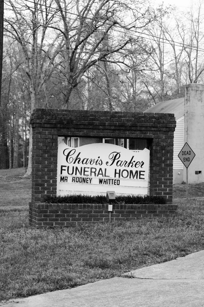 A black and white photo of a brick‑framed roadside sign reading “Chavis Parker Funeral Home” with a changeable‑letter panel below stating “Mr Rodney Whitted.” The sign sits on a grassy lawn by a driveway; behind it are bare winter trees, a “Dead End” street sign, power lines overhead, and part of a house’s exterior wall