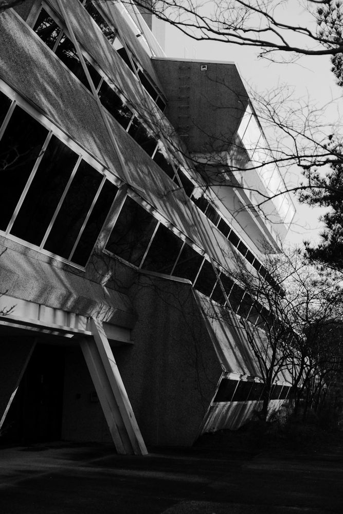 A black and white photo of the Burroughs Wellcome building in Research Triangle Park. Now demolished, it is an angular concrete structure with large windows spanning its length. The structure leans sharply to the left, creating a dramatic diagonal perspective. Leafless tree branches in the foreground cast shadows on the building, enhancing the stark, eerie atmosphere. The scene appears to be taken during winter or early spring, with a cold and quiet mood