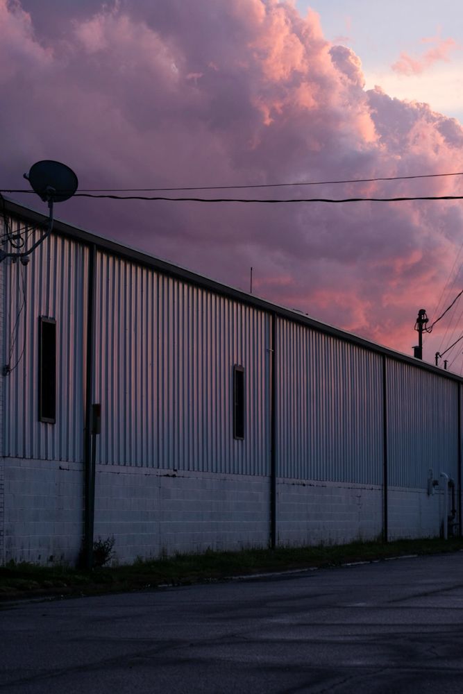 A photo of a long, corrugated metal warehouse building at dusk, with dramatic pink and purple storm clouds filling the sky above. Power lines stretch across the top of the frame, and a satellite dish is mounted on the corner of the building. The building's siding reflects the colors of the sunset, casting soft purples and warm pinks on its surface. The street in the foreground is wet and slightly reflective.