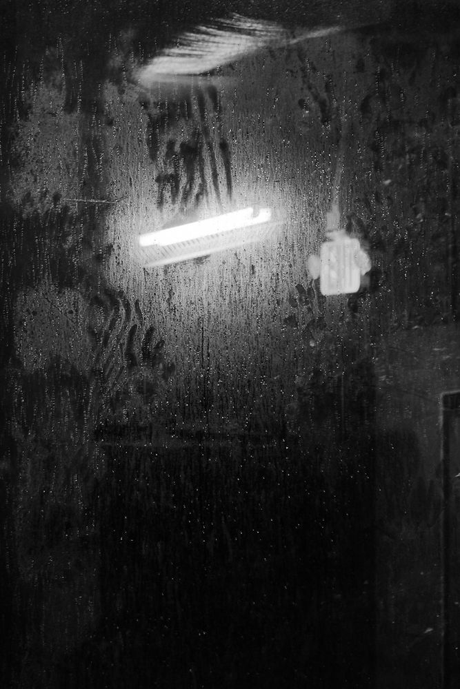 A black and white photo of a fogged-up glass surface, covered in condensation and smudged handprints. A fluorescent ceiling light and an electrical outlet are faintly reflected in the glass, giving the scene an eerie, moody atmosphere
