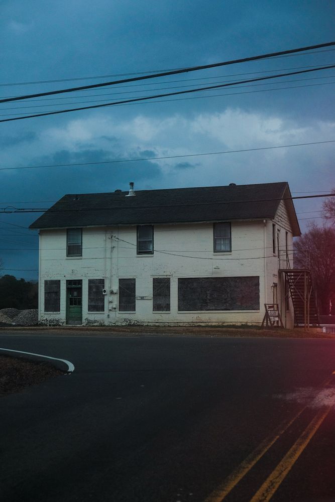 A photo of a two-story, boarded-up white building at dusk, situated at a road intersection. The windows on the ground floor are covered with weathered plywood, and the upper windows are bare but appear aged. A narrow metal staircase leads to a side entrance on the second floor. Power lines cross the sky, which is overcast with dark blue storm clouds. The scene is dimly lit, with a faint red light leak or lens flare in the bottom right corner.