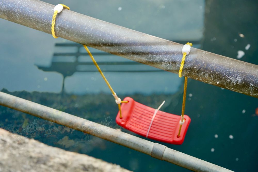 A red swing hangs from a railroad bridge over a river reflecting and contrasting with another photo of a park bench and tree superimposed over it.