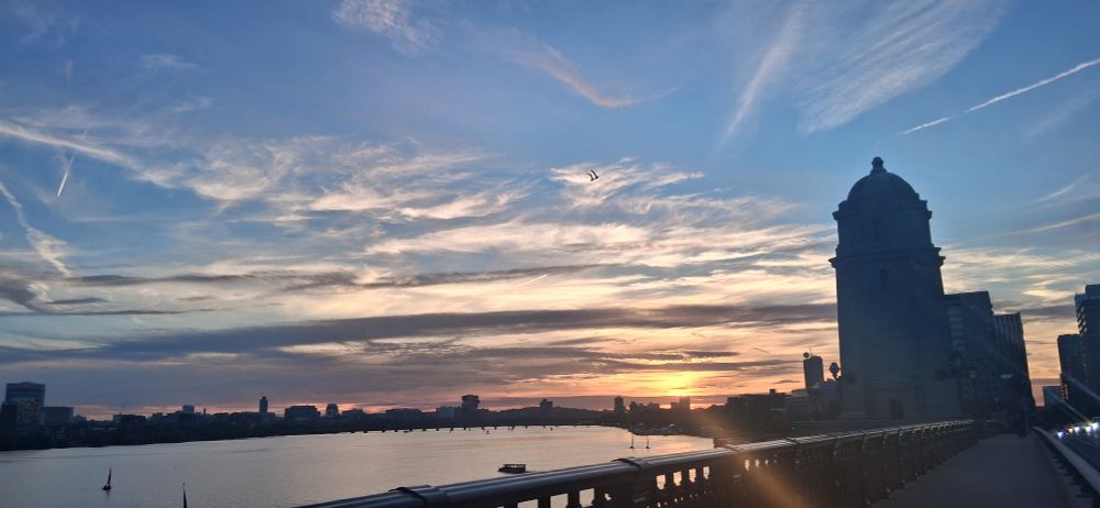 A sunset over a river with a glowing sun and hues of pink orange purple going all the way to light blue threres a city in the background 