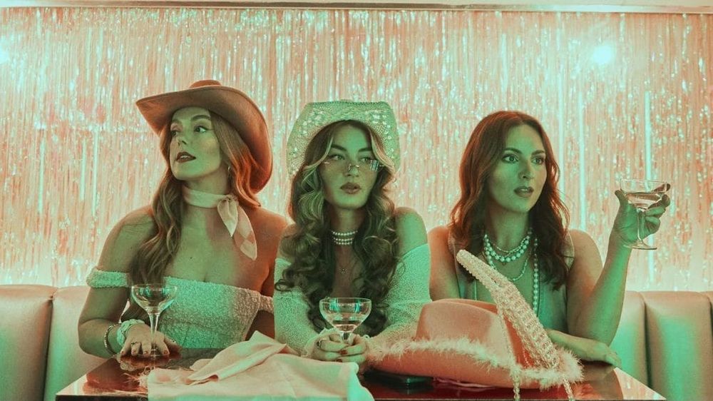 The three members of female pop-country trio, Remember Monday, sitting at a table against a glittering backdrop, wearing cowgirl hats and holding cocktail glasses