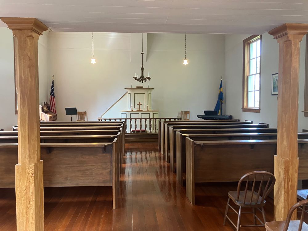 The interior of the chapel, which has bare white walls and plain glass windows. Wooden pews with an aisle down the middle face a raised alter and a higher pulpit behind it. 