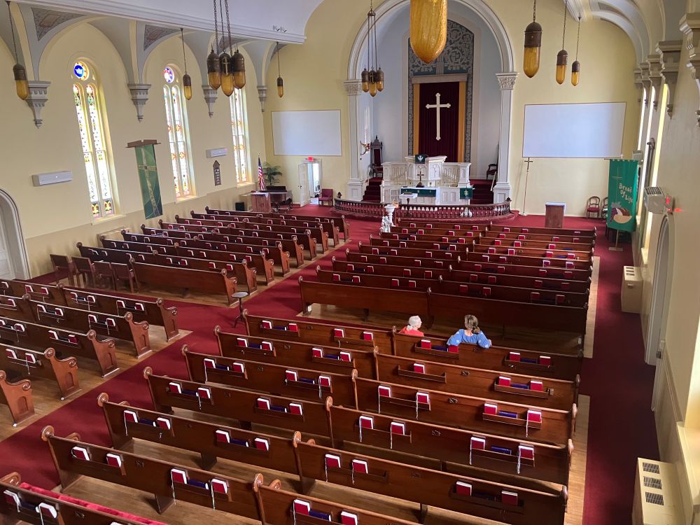 The interior of the church with row after row of pews facing a white altar. The other walls are yellow, and the windows have stained glass (probably not original). 