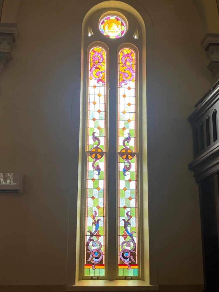 A stained glass window. Two long panels with a circular window above.  This window shows a beehive above.