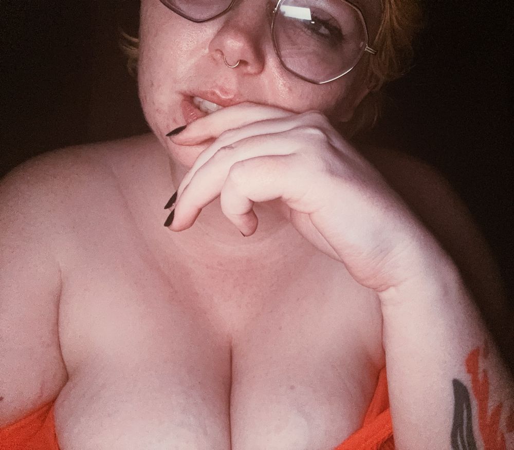a white nonbinary person in glasses with short orange hair flirts with the camera. they are in an orange bra with the straps pulled down to expose their chest and have a hand to their mouth in a seductive pose