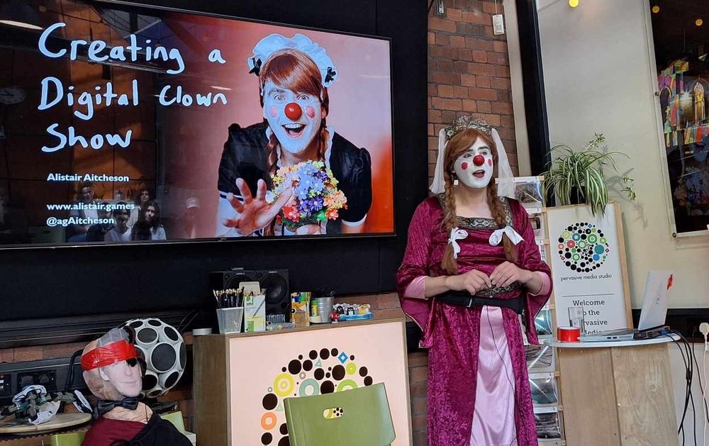 Alistair as clown Juliet, in front of a slide showing him as clown Cafetière (the French maid), with title "Creating a Digital Clown Show". Pierce Brosnan, in the corner, has a feather gaffer taped over his eye in a way that makes him look like Solid Snake