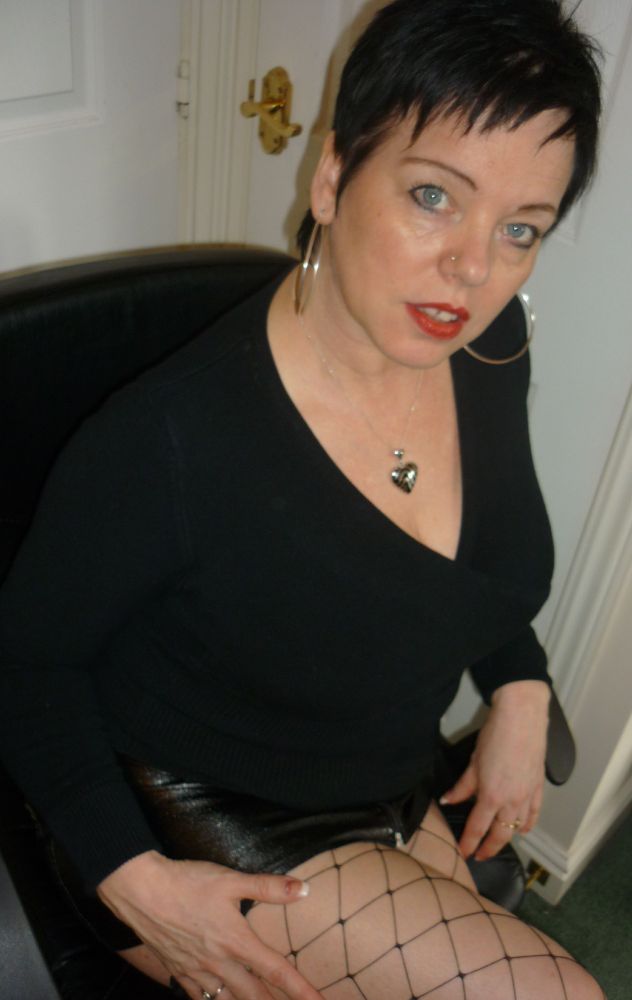 A photo of mature, busty, Donna X. A throwback pic from when I had black short crop "pixie" haircut. I am wearing a black foldover crop top and a black leather skirt with fence net stockings.