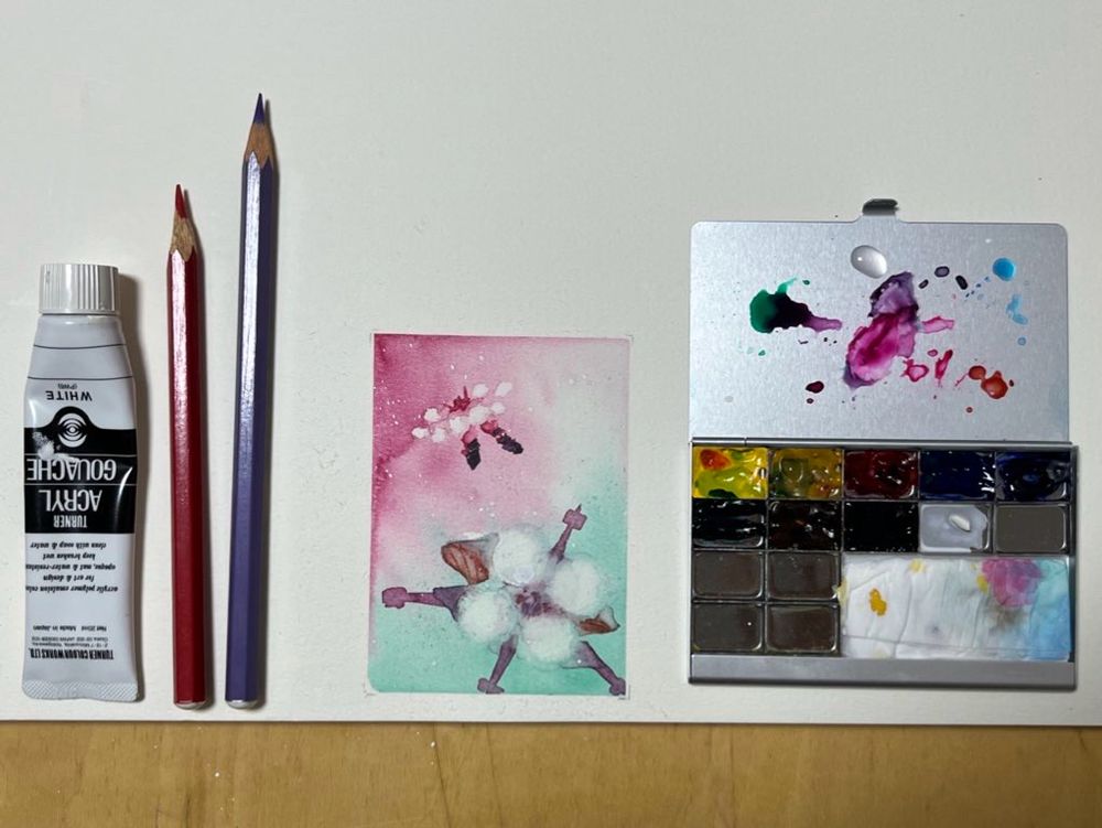 水彩画の周りに画材や色鉛筆、白いアクリル絵の具が並べられている。