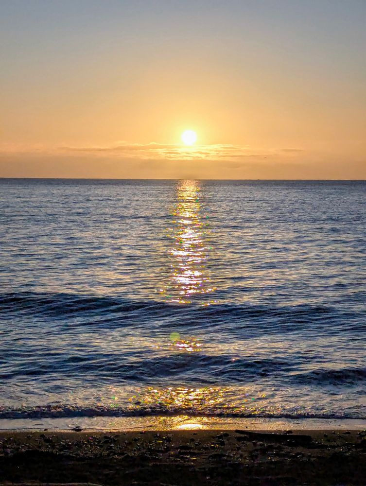 A bright yellow sun breaks through the clouds while casting vibrant yellow light upon the gently rippling ocean waves of the Esquimalt Lagoon Beach 
