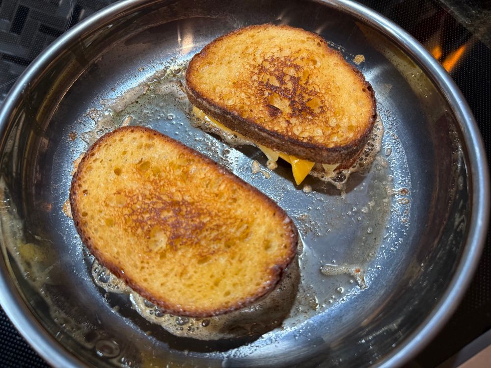 Two small golden grilled cheese sandwiches cooking in a silvery skillet. There’s a lot of oil in the bottom of the pan just from the melted butternaise!