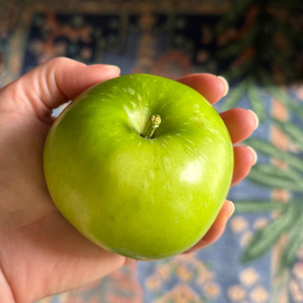 A shiny bright green palm-sized apple held in a hand over a colorful carpet 