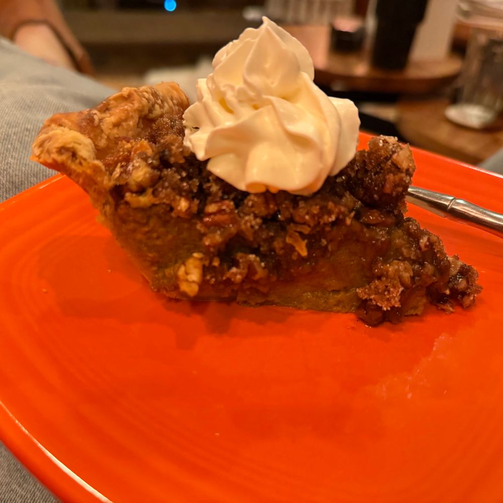 A slice of combination pecan pumpkin pie on a bright orange plate with a spiral of spray whipped cream on top 