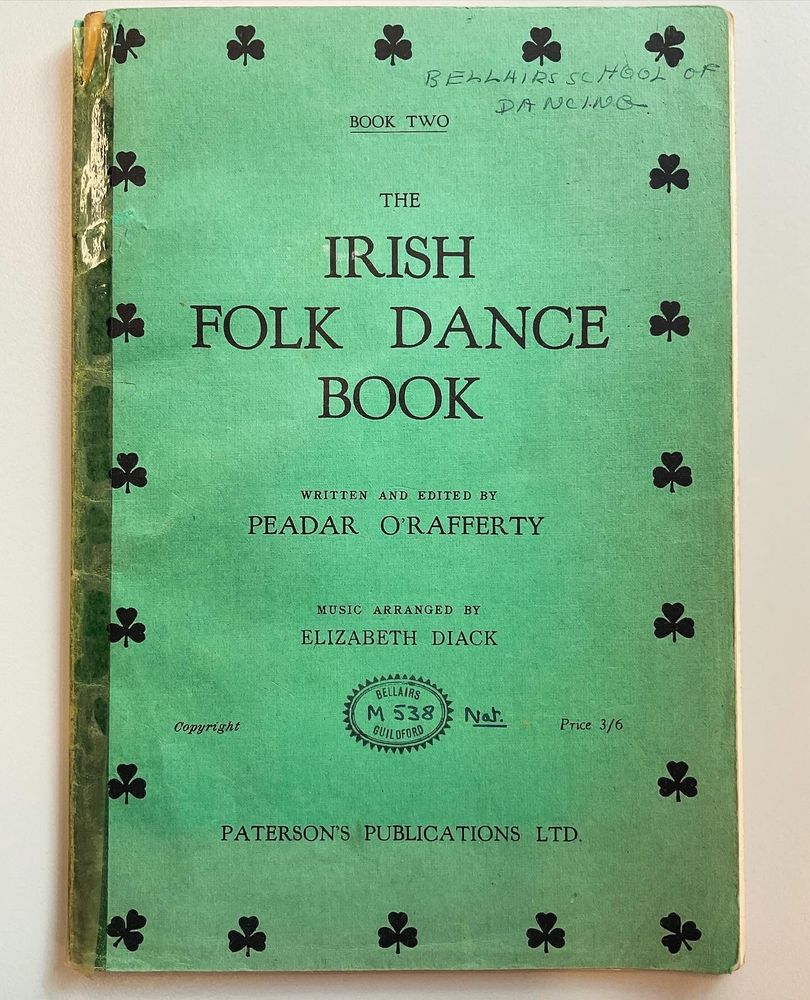 A battered emerald green book of Irish Folk Dances, written and arranged by Peadar O'Rafferty. There is a jaunty pattern of shamrocks around the edge, and along the top is written 'Bellairs School of Dancing'. 
The spine is held together with aging sticky tape, one of the things us archivists really dislike as it leaves a residue, but it shows how much use this book got during its life.