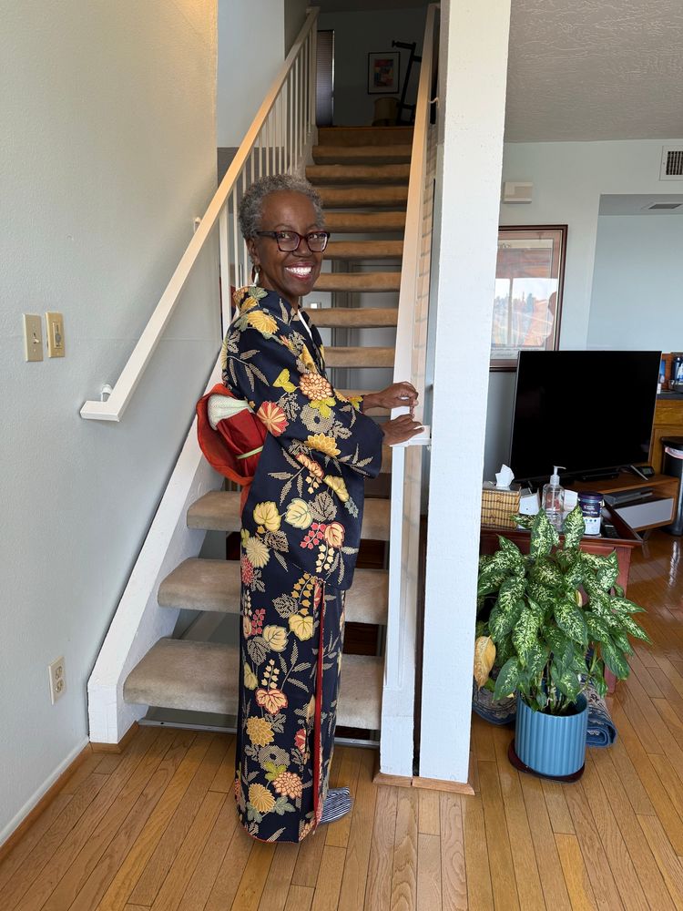 My beautiful mother by the stairs posing in my wife’s kimono passed down through her family. The kimono is black or navy with many gold and red flowers and a red obi. She has earrings, glasses and a big smile 