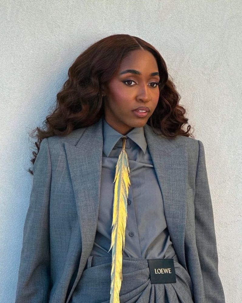 Ayo Edeberi at a photo shoot for the Golden Globes wearing a grey blazer, grey slacks and a grey shirt and black dress shoes with a gold feather tie 