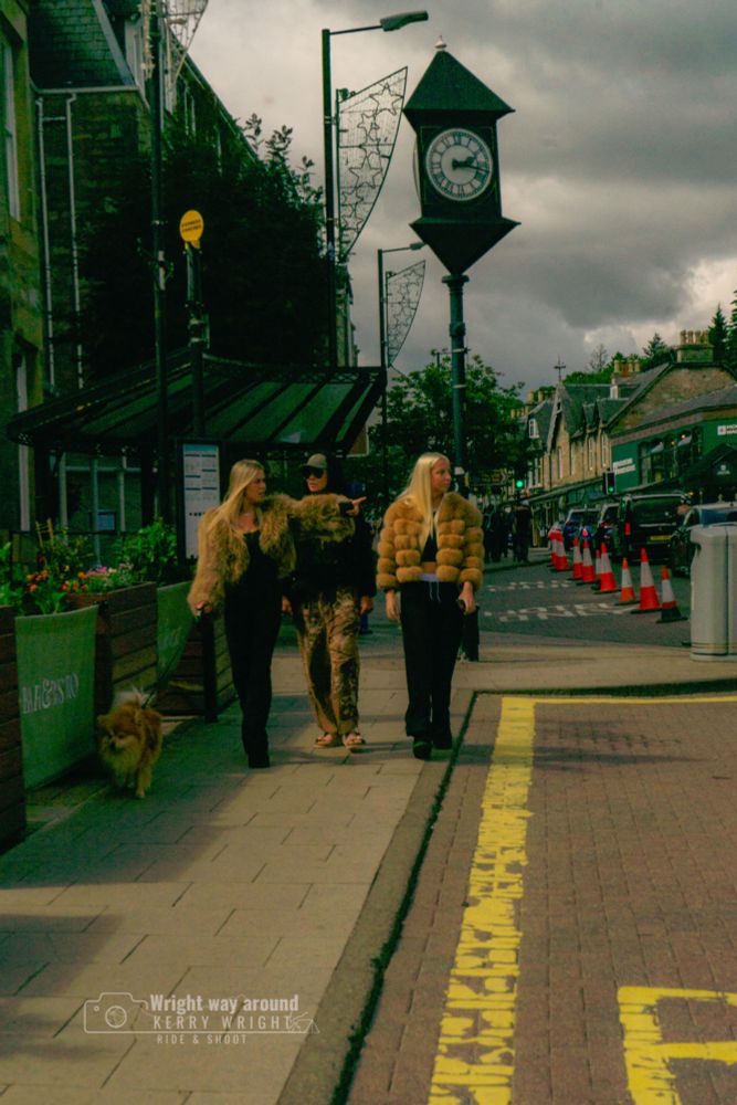 Two women in fur coats walking a dog on a busy street, captured candidly. The photo has a warm, grainy tone that gives it a 1970s film vibe.