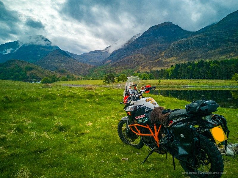 A KTM 1190 motorcycle parked in the scenic village of Kinlochleven. The bike is rugged and adventure-ready, standing out with its distinctive orange and black design. The surrounding landscape features clear skies and the dramatic, mountainous terrain of the Scottish Highlands, evoking a sense of freedom and adventure. The image captures a moment of connection with the bike, perhaps waiting for the right part to fix or enhance it.