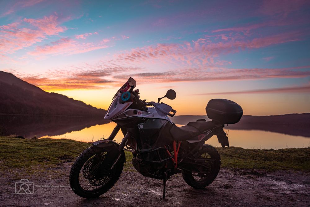 A motorcycle parked near the edge of Loch Rannoch, with the bike silhouetted against the tranquil, sunset-lit water reflecting the soft glow of the sky.
