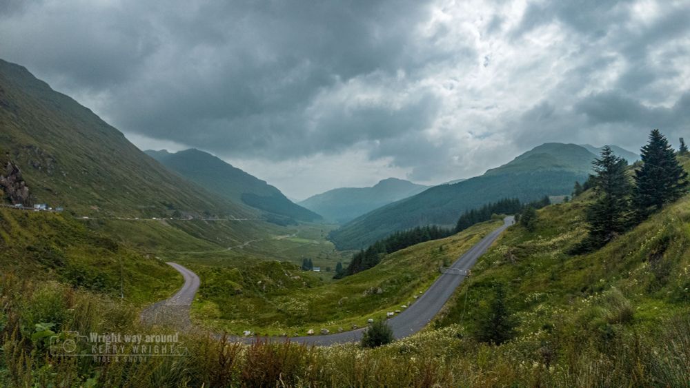 View of The Rest and Be Thankful, a historic mountain pass in the Arrochar Alps of the Scottish Highlands. The A83 road winds through steep glens surrounded by lush green hills and rugged peaks. Low clouds cling to the slopes, adding drama to the remote, misty landscape. The pass sits at the junction of Glen Kinglas and Glen Croe and offers panoramic views of the surrounding highlands. A stone marker, built by soldiers in the 1750s, reads “Rest and Be Thankful”—named because weary travellers and military road builders would pause here after the steep climb. The area has a long history of landslides due to unstable slopes, with major events every few years. Ongoing roadworks and landslide barriers are visible, highlighting the challenges of maintaining this vital route.