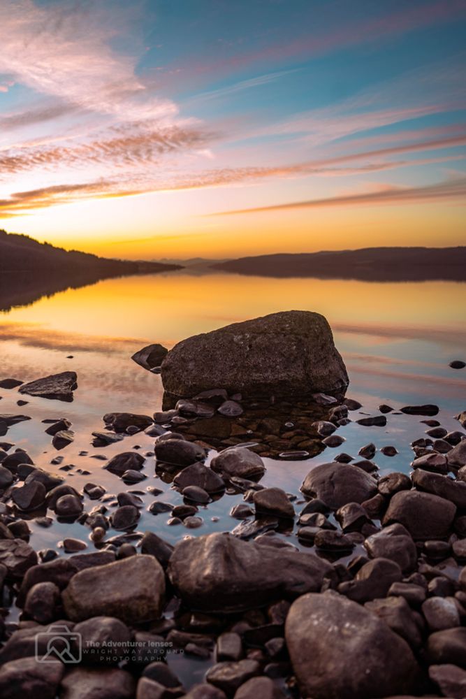 Sunset over Loch Rannoch in Perthshire, Scotland. The calm water reflects the sky like a mirror, with warm tones of orange, pink, and purple stretching across the horizon and mirrored perfectly on the loch’s surface.
