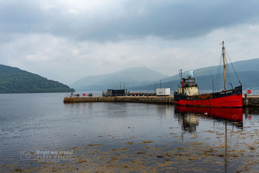 A moody view of Inveraray Pier on the shores of Loch Fyne, with the historic red and cream puffer boat Vital Spark moored on the right. Calm, seaweed-speckled waters reflect the boat and the surrounding misty Highlands. In the background, forested hills fade into blue-grey tones under a heavy, clouded sky. Vital Spark is a famous Clyde puffer, a type of cargo ship once essential to Scottish coastal trade in the 19th and early 20th centuries. This particular vessel is named after the fictional boat from Neil Munro’s beloved Para Handy stories, which humorously chronicled life aboard a West Highland steamer. Loch Fyne, Scotland’s longest sea loch, was historically vital for fishing, trade, and naval routes, and today it’s known for its oysters and scenic beauty. The scene blends history, folklore, and Highland atmosphere in one peaceful moment.