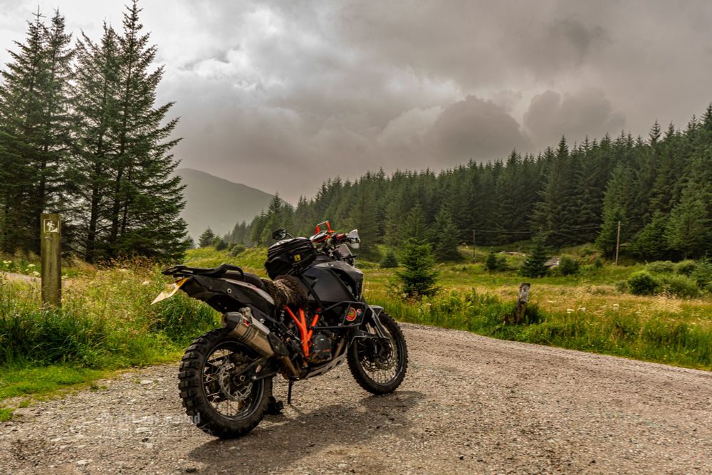 A photo of my motorbike parked along the quiet B828 road in the Scottish Highlands. Sunlight breaks through the clouds, casting a golden glow over the bike and the surrounding moorland. The narrow, winding single-track road stretches into the distance, flanked by rugged hills, patches of wild grass, and heather. The B828 is a remote route near Loch Goil, offering dramatic Highland views with very little traffic—perfect for solo riding and scenic exploration. The sunlight catching the bike highlights its details and upgrades, making it stand out against the natural, earthy backdrop.