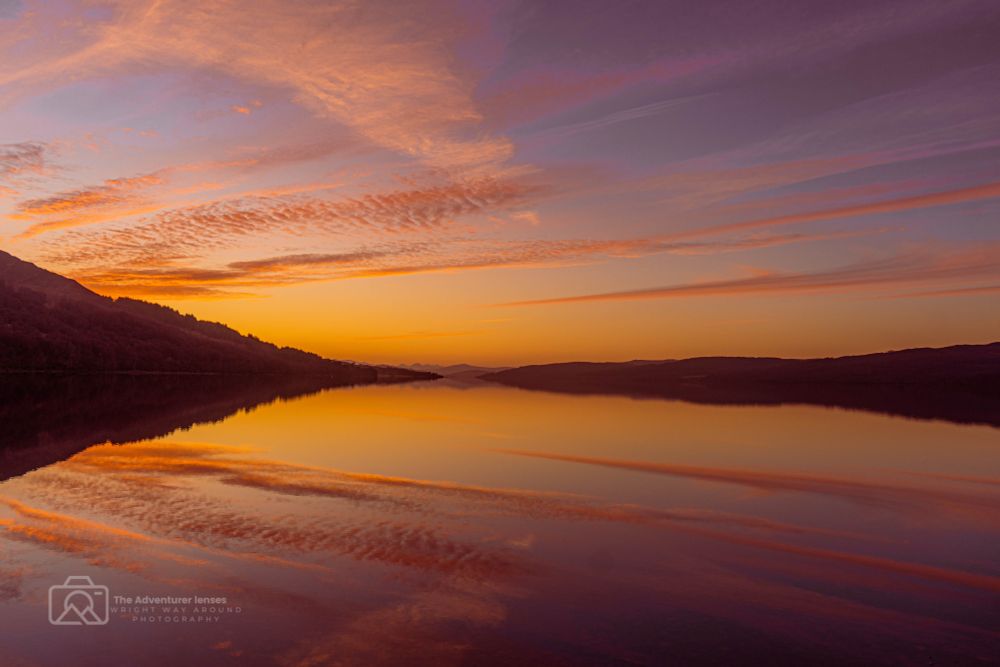 Still waters of Loch Rannoch at sunset, perfectly mirroring the glowing sky with soft hues of orange, pink, and purple stretching across the horizon.