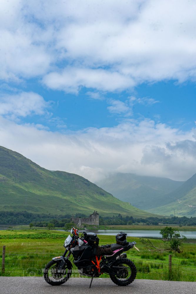 historic ruins of Kilchurn Castle in the background, nestled beside a tranquil Scottish loch and framed by misty hills. Overcast sky adds to the dramatic Highland atmosphere.