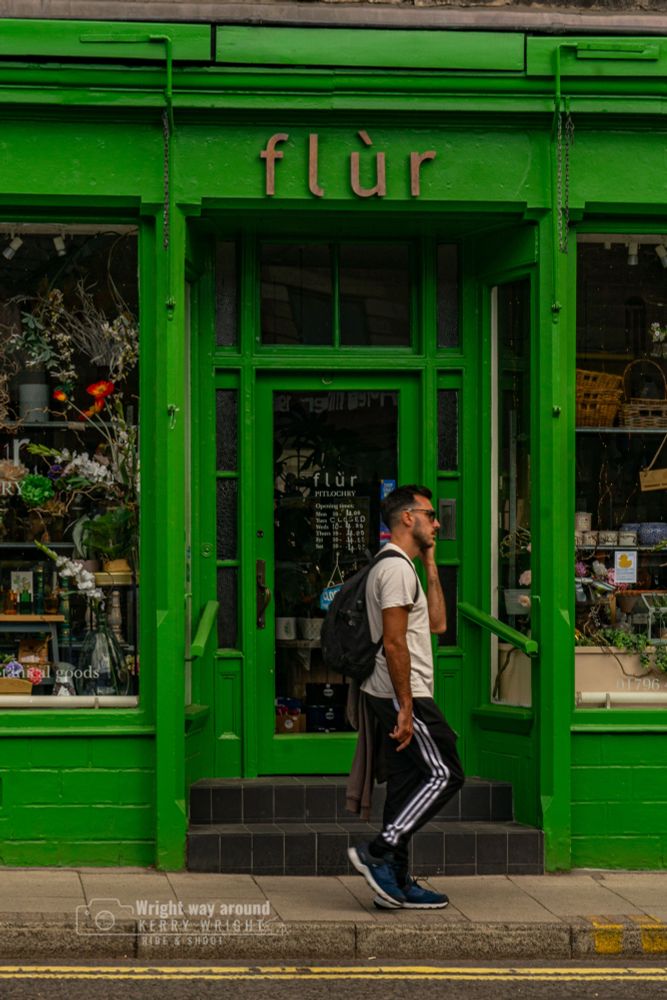 A man walking past a green flower shop while talking on his phone, perfectly framed by the closed door. A candid street moment with a vintage, cinematic feel.