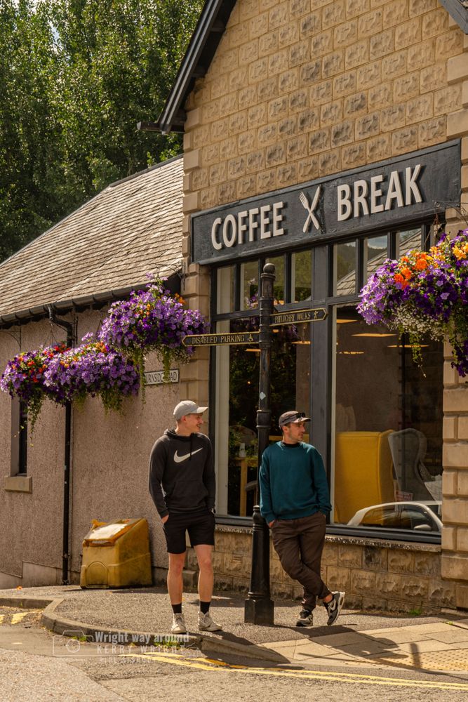 Two men on a street corner, one leaning casually against a tall signpost with directional signs for visitors. Behind them, a café with "Caffè Breaks" signage adds to the urban backdrop. The scene captures a relaxed moment in the middle of town, blending human interaction with everyday city life. Natural light highlights the textures of the street and the quiet atmosphere.