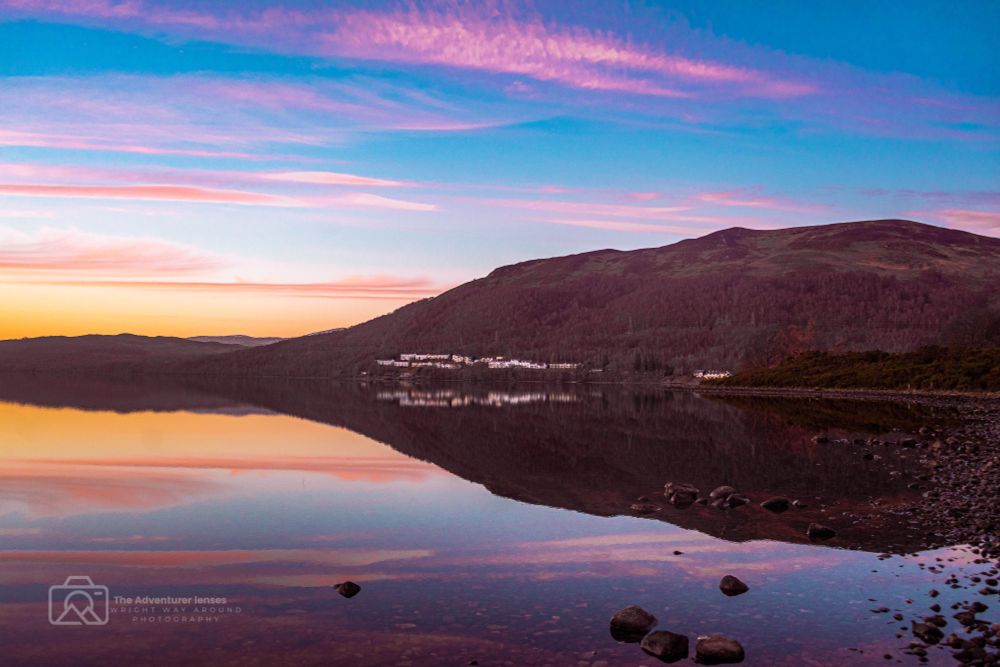 Rannoch Resort nestled on the shore of Loch Rannoch, surrounded by trees and hills, with the calm loch and pastel sunset sky creating a peaceful Highland scene."