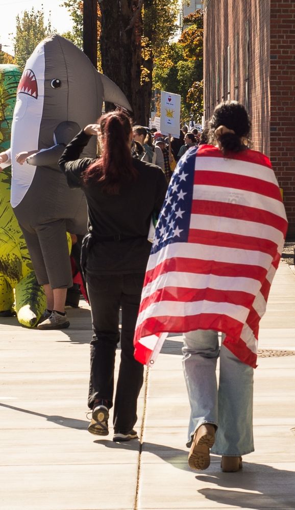 No Kings protest Benton County OR 10-18-25. A young person is pictures wrapped in the American flag walking down the street with a crowd in front of them.