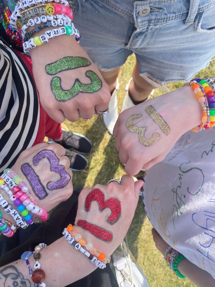 Four fans of Taylor Swift have their hands in a fist held out to each other as they stand in a circle. Each one displays a different color glittered number 13: one each in red, gold, purple, and green. Every wrist is adorned with lovingly made friendship bracelets to trade at the Eras Tour. This is girlhood. 🫶🏻