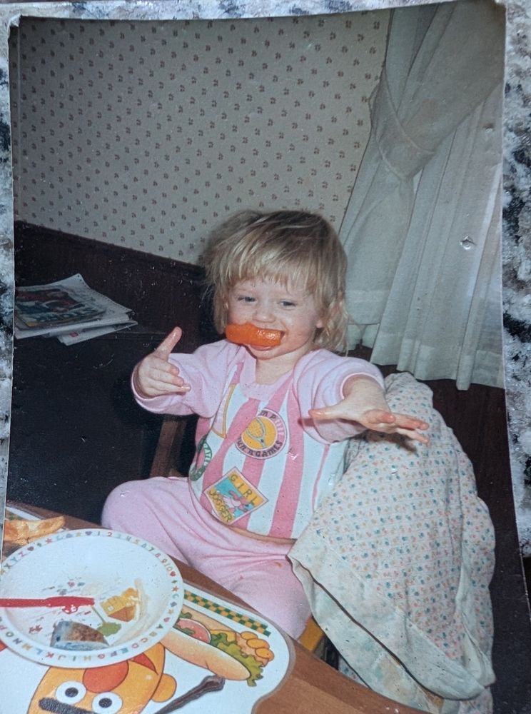 A toddler with blonde hair reaches her arms toward the camera. She has a slice of something orange in her mouth, maybe an orange bell pepper or an orange slice and she is grinning. She is wearing pink pants and a white and pink shirt. On the arm of her chair is a light pink blanket with satin trim. She is sitting in a high chair at a table. On the table is a sesame Street placement featuring Bert with a hot dog. On top of the placement is her plate, and it looks like the orange slice in her mouth is the last bite of her meal. 