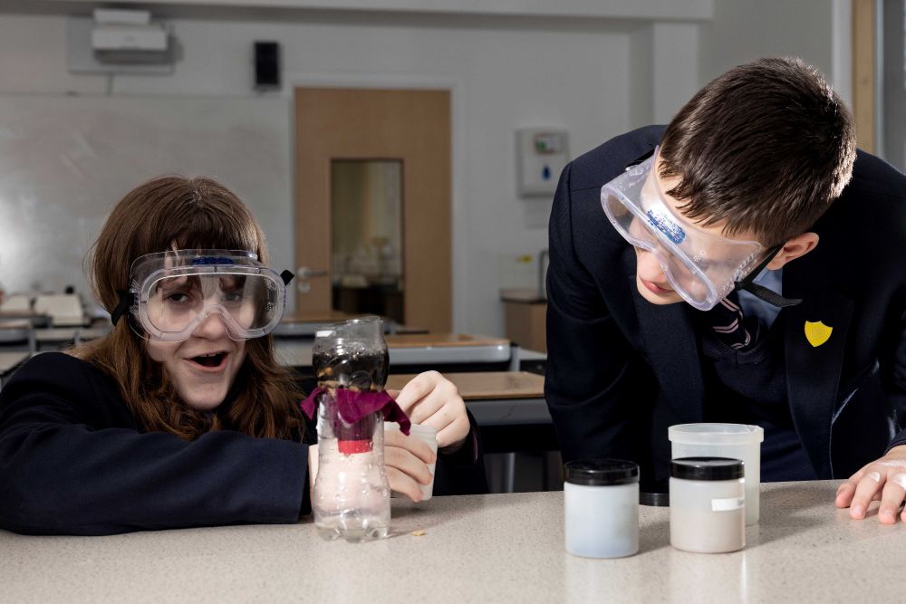 2 school students wearing protective equipment conducting a science experiment with beakers and chemicals