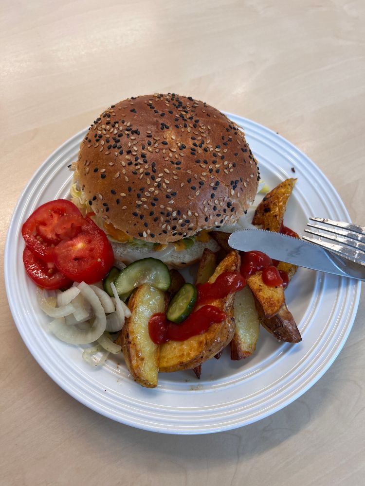 Hamburger med stekte poteter, pickles og tomat på en hvit tallerken.