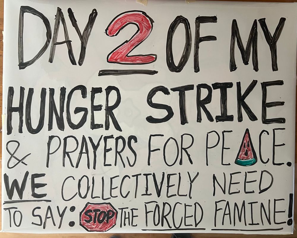 Wearable Whiteboard sign reading in bold black and red letters: “Day 2 of my hunger, strike and prayers for peace. We collectively need to say: STOP the forced famine!”