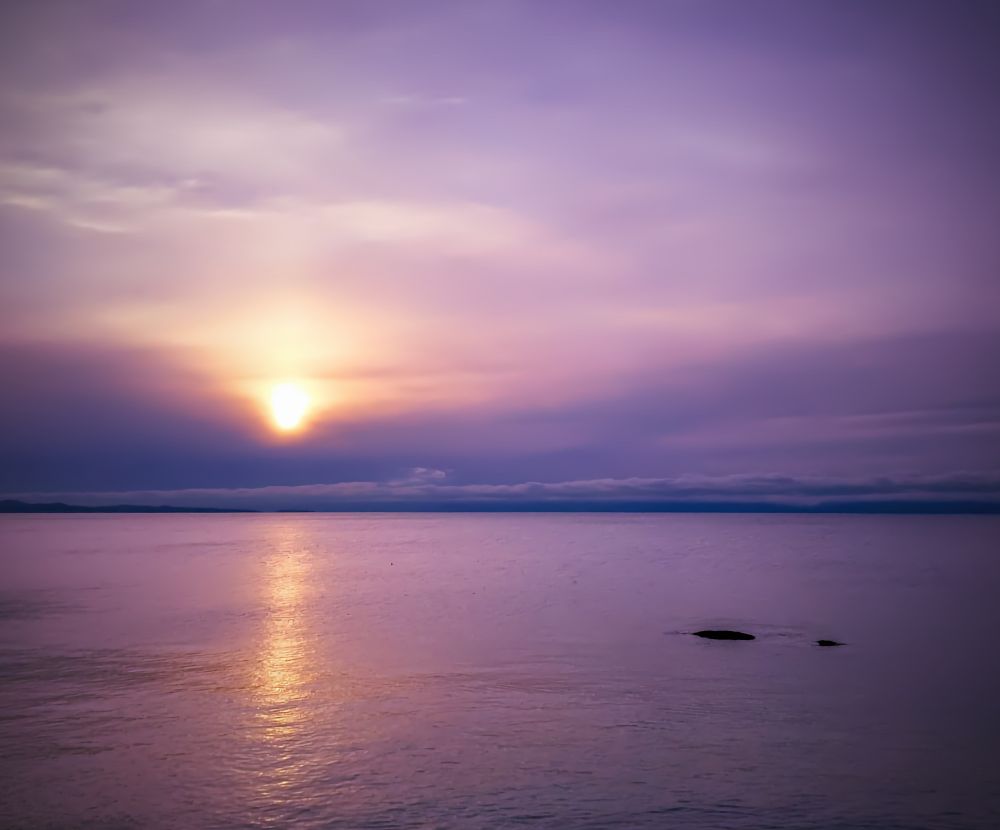 The sun slowly sinking across the pacific ocean. Long streaky clouds hang gently over a distant land mass. The sky is almost purple save for a golden streak in the calm water
