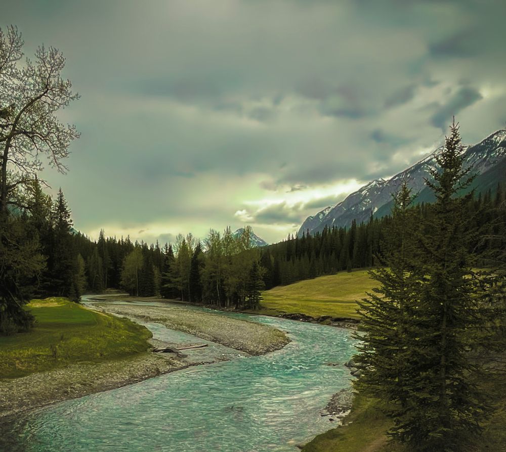 A blue/green river winding its way through a tree lined valley at the base of a glacial mountain. Clouds cling to the mountain tops with a hint of brightness beyond.