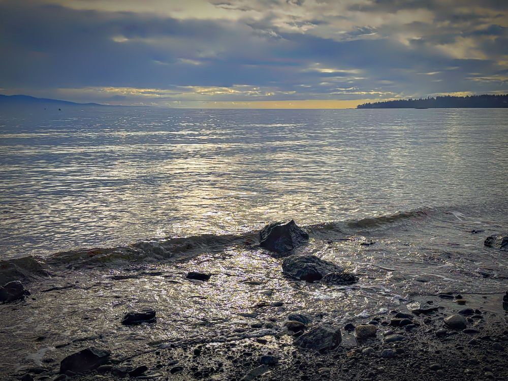 A rocky shore with calm water. A small wave begins its break on a jagged rock at the shoreline. The sky is overcast and low, hanging above the water. In the distance a couple of land masses and a sliver of brightness in yellow tones.  